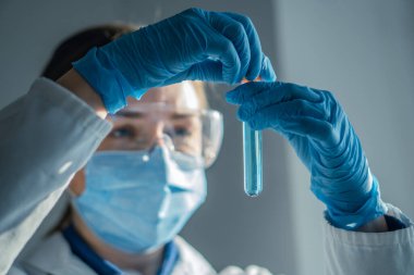 Koruyucu giyimde yetenekli bir laboratuar bilimcisi canlı bir mavi sıvıyı test tüpünde titizlikle inceliyor ve bilimsel araştırmalarda güvenliğe ve hassasiyete olan bağlılıklarını gösteriyor.