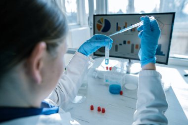 Beyaz önlüklü ve mavi eldivenli yetenekli bir laboratuvar çalışanı, örnekleri titizlikle pipetle hazırlıyor. Monitördeki grafikler ve bilgi grafikleri bilimsel çalışmaların önemini vurguluyor.