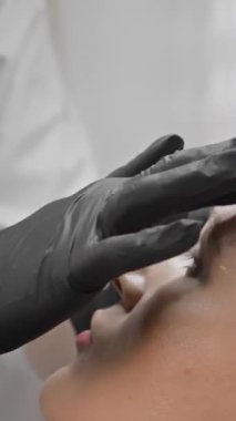 Siyah eldivenli bir güzellik uzmanı, spa ya da klinik ortamında hastaların alnına cilt bakımı yapıyor. Saç bandı takan hasta profesyonel bir çevrede.