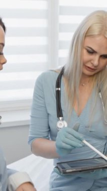 Bir doktor, modern bir klinikte bir hastayla görüşürken tablet kullanır. Tıbbi tedavi seçeneklerini tartışıyor gibi görünüyorlar, hasta merkezli bakım ve dijital sağlık teknolojisini vurguluyorlar.
