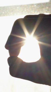 Bir dizi çerçeve çeşitli şekiller oluşturan bir el görüntüsü, parlak ışıklı bir pencereye karşı siluet şeklinde. Parlak güneş ışığı parmaklarının arasından parlar ve çarpıcı bir halo etkisi yaratır..