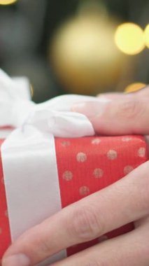 Beyaz kurdeleli canlı kırmızı bir hediye kutusu renkli süsler ve sıcak ışıklarla süslenmiş neşeli bir Noel ağacının önünde gururla sunuluyor ve neşeli bir atmosfer yaratıyor.