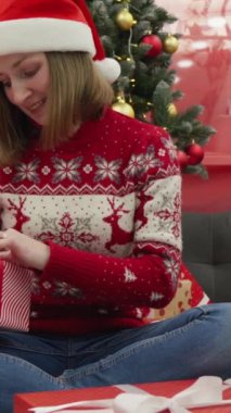 Neşeli bir tatil sahnesinde, Noel Baba şapkası ve şen süveter giyen genç bir kadın, güzel dekore edilmiş bir ağacın altında neşe içinde Noel hediyelerini açıyor ve mevsimlerin büyüsünü arttırıyor.