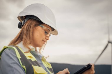 Kendini güvenlik donanımına adamış bir kadın mühendis rüzgar çiftliğindeki tabletindeki performans verilerini inceleyerek sürdürülebilirlik ve yenilikçi yenilenebilir enerji çözümlerine olan bağlılığını gösteriyor.