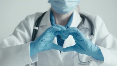 Maske, mavi eldiven ve steteskop takan bir tıp uzmanının eldivenli elleriyle kalp şeklini oluşturduğu yakın plan bir video. Bu sağlık hizmetlerinde bakım, bağlılık ve bağlılığı sembolize eder.