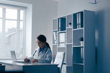 Parlak ve modern bir ofiste, kendini adamış bir kadın doktor hasta bilgilerini bilgisayarından inceler ve notlar alır, hepsi de sakin, sakin bir ortamda, sağlık ve profesyonel bakımı teşvik eder.