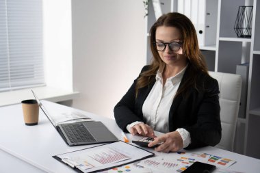 Resmi giyinmiş bir iş kadını modern ofis masasında özenle çalışıyor. Akıllı telefonundaki verileri analiz ediyor ve bilgisayarındaki raporları inceliyor. Verimliliğe olan bağlılığını gösteriyor.