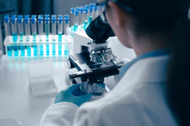 Laboratuvar önlüğü giymiş odaklanmış bir bilim adamı mikroskop altında örnekleri inceliyor, modern keşifler için modern bir araştırma ortamında test tüpleri ve gelişmiş ekipmanlarla çevrili.