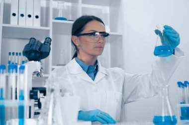Parlak, düzenli bir laboratuvarda, beyaz laboratuvar önlüğü ve gözlüklü bir kadın, bilimsel bilgi ve sağlık hizmetlerine olan katkılarını ve araştırmalarındaki rolünü vurgulayan canlı mavi bir sıvı şişesini inceliyor.