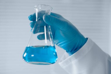 Steril bir laboratuvarda, laboratuvar önlüğü ve mavi eldivenli kendini adamış bir bilim adamı canlı mavi bir sıvıyla Erlenmeyer şişesini tutuyor ve bilimsel araştırmaların büyüleyici dünyasını vurguluyor.