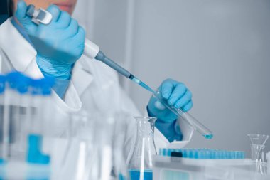 Beyaz önlüklü ve mavi eldivenli bir laboratuvar teknisyeni sıvıyı test tüpüne aktarmak için dikkatlice pipet kullanıyor..