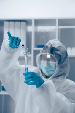 Koruyucu giysiler içindeki bir bilim adamı, steril bir laboratuvar ortamında deneysel teknikler sergileyerek test tüpünde mavi bir sıvıyı dikkatle gözlemler.