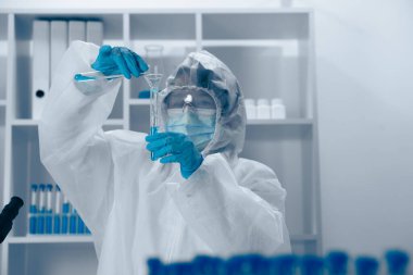 Temizlenmiş bir laboratuvarda, koruyucu giysi içinde kendini adamış bir araştırmacı test tüplerine canlı bir sıvı dökerek gerekli deneyleri gerçekleştirmede hassasiyet ve beceri gösteriyor.
