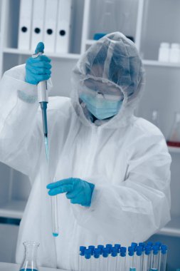 Koruyucu giysiler içindeki maskeli bir bilim adamı, steril bir araştırma ortamı sunan deney tüpleri ve laboratuvar ekipmanlarıyla çevrili bir pipet kullanarak sıvı kullanımını dikkatlice gerçekleştiriyor..