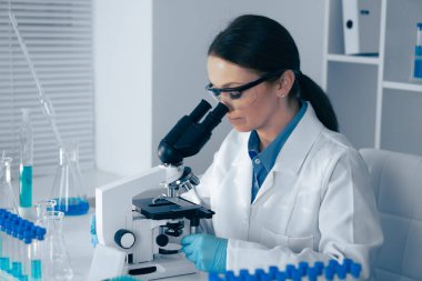Gıcır gıcır laboratuar önlüğü giymiş odaklanmış bir kadın bilim adamı, bilimsel araştırmalara olan bağlılığını gösteren cam eşyalar ve mavi test tüpleriyle dolu modern bir laboratuvarda, mikroskop altında örnekleri inceliyor.