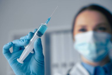 Eldivenli maskeli bir sağlık görevlisi elinde mavi sıvıyla bir şırınga tutuyor. Herkesin güvenliği ve hijyeni sağlamak için katı sağlık protokollerini takip ederken aşı yapmaya hazırlanıyor.
