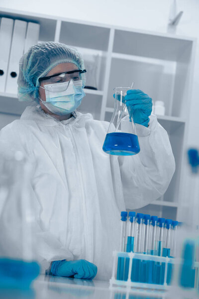 Сосредоточенный ученый в СИЗ исследует фляжку яркой голубой жидкости в современной лаборатории, демонстрируя точность и безопасность, необходимые в современных биохимических и биотехнологических исследованиях