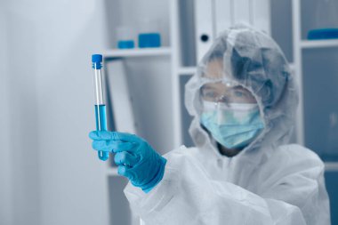 Koruyucu giysiler içindeki titiz bir laboratuar bilimcisi test tüpündeki canlı mavi sıvıyı inceliyor. Steril bir laboratuvar ortamında güvenlik protokollerine ve hassasiyete güçlü bir bağlılığı yansıtıyor.