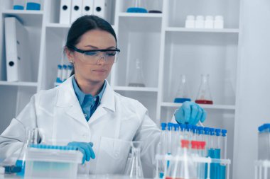 Beyaz laboratuvar önlüğü ve koruyucu gözlüklü kendini adamış bir kadın bilim adamı modern bir laboratuvarda renkli test tüplerini analiz ediyor ve araştırmaya ve bilimsel gelişime olan bağlılığını gösteriyor.