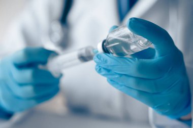 Klinik bir ortamda, mavi eldivenli bir sağlık görevlisi şırıngadan şırıngaya sıvı çekerek aşı hazırlama ve hastanın güvenliğini sağlama sürecini vurguluyor.