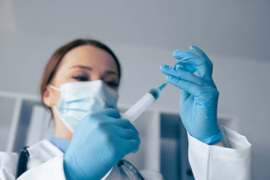 Beyaz önlüklü ve koruyucu teçhizatlı bir sağlık uzmanı, aşı şırıngası hazırlar ve optimum hasta bakımı için kontrollü bir klinik ortamında güvenlik ve hassasiyete dikkat çeker.