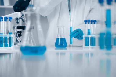 Laboratuvar önlüklü bir bilim adamı titizlikle test tüpleri ve canlı mavi sıvı içeren bir şişeyle çalışır. Modern bir laboratuvarda bilimsel deneylerin hassasiyetini gösterir..