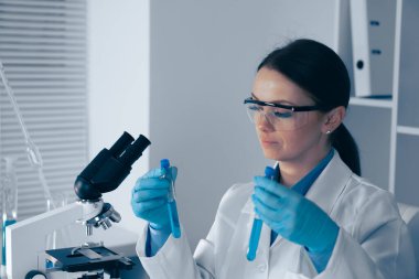 Bilimsel bir araştırma laboratuvarında kendini adamış beyaz önlüklü bir bilim adamı mikroskobun yanındaki iki test tüpünü inceliyor. Anahtar kelime analizi, biyokimya, biyomedikal...