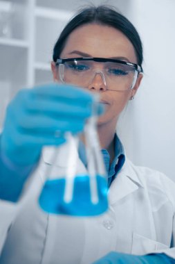 Organize bir laboratuvarda, laboratuvar önlüğü ve güvenlik gözlüğü takmış odaklanmış bir bilim adamı canlı mavi bir sıvıyı inceleyerek bilimsel araştırmalarda hassasiyetin önemini vurguluyor.