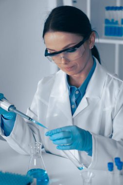 Modern bir laboratuvarda, laboratuvar önlüğü ve güvenlik gözlüğü takmış odaklanmış bir kadın bilim adamı sıvıyı bir şişeye aktarmak için ustalıkla pipet kullanır ve biyoteknoloji ve kimya konusundaki yeteneklerini gösterir.