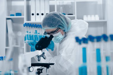 Koruyucu ekipman kullanan kendini adamış bir bilim adamı, bilimsel araştırmalar için tasarlanmış steril bir araştırma ortamında gelişmiş teknikler sergileyen test tüplerini mikroskop altında inceliyor.