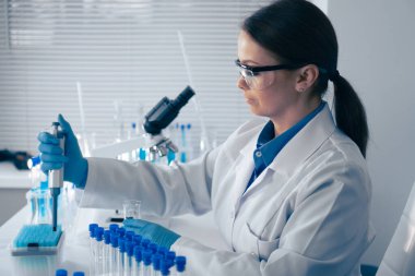 Parlak bir laboratuvarda, beyaz önlüklü kendini adamış bir kadın bilim adamı önemli araştırması için gerekli ekipman ve test tüpleri ile çevrili bir mikropipetle sıvıları titizlikle ölçer.