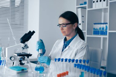 Beyaz önlüklü bir bilimadamı, temel bilimsel araştırmalar için gerekli aletler ve test tüpleriyle çevrili sıvı çözeltileri dikkatle ölçer.