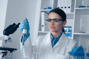 Laboratuvar önlüğü ve güvenlik gözlüğü takan yetenekli bir kadın bilim adamı sıvıyı nakletmek için mikro pipet kullanır, araştırma ve hassasiyet ve profesyonelliğin önemi üzerine uzmanlığını sergiler.