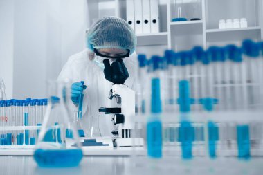 Tutkulu bir bilim adamı deney tüpleriyle dolu modern bir laboratuvarda mikroskop altında numuneleri inceliyor ve ilginç özelliklerini vurgulayan canlı mavi sıvılar içeren gelişmiş aletleri inceliyor.