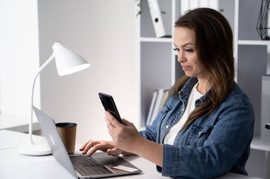 Kendini işine adamış profesyonel bir kadın şık bir ofiste dizüstü bilgisayar ve akıllı telefon kullanarak rutinindeki teknoloji ve verimliliğin karışımını sergiliyor.