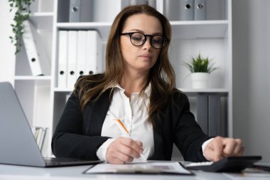 Gözlük takan odaklanmış bir kadın titizlikle modern ofis masasındaki verileri analiz ediyor, özenle notlar yazıyor ve hesap makinesi kullanıyor.