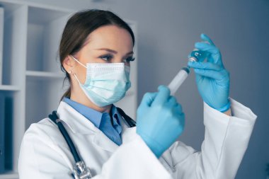 Koruyucu maske takan ve mavi eldiven takan bir sağlık uzmanı titizlikle bir şişeden aşı hazırlıyor. Bu da hasta bakımı ve güvenlik protokollerinde gayret gösterdiğini gösteriyor.