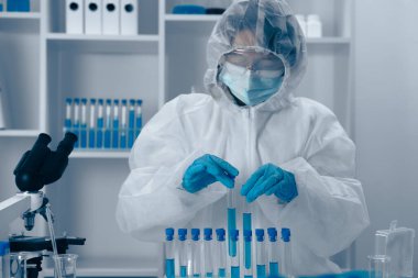 Koruyucu giysi giyen kendini adamış bir bilim adamı, sıkı güvenlik protokollerini takip ederek ve araştırmalardaki detaylara olağanüstü özen göstererek refah dolu bir laboratuvarda test tüpleriyle dikkatlice ilgilenir.
