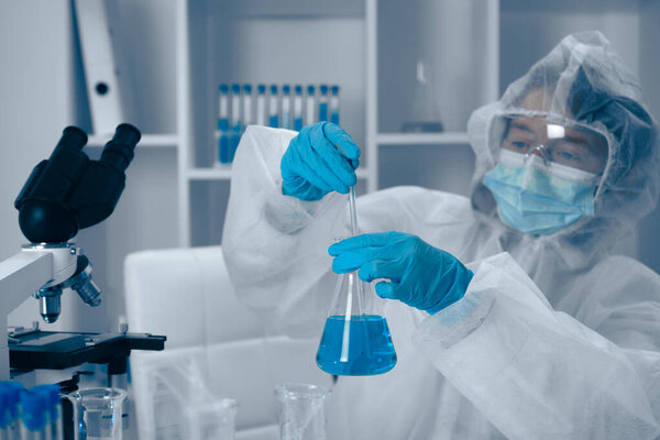 In a stateoftheart modern laboratory, a dedicated scientist wearing protective gear is meticulously measuring a vivid blue liquid from a conical flask with utmost precision and care