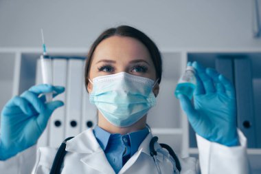 Klinik bir ortamda, beyaz önlük, eldiven ve koruyucu yüz maskesi takan bir sağlık uzmanı hayati bir enjeksiyona hazırlanan bir şırınganın yanında güvenilir bir şekilde bir şırınga tutuyor.