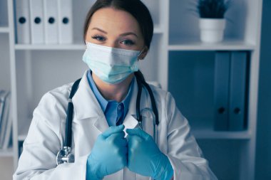 Beyaz önlüklü, maskeli ve eldivenli kendini adamış bir kadın sağlık uzmanı bir hastayla meşgul oluyor. Klinik bir ortamda güvenliğe ve kaliteli bakıma olan bağlılığını gösteriyor.