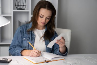 Kendini işine adamış bir kadın, finansal makbuzları titizlikle inceliyor ve modern masasındaki bir deftere notlar yazıyor. İşlerinde başarıyı arttıran verimli bir ortam yaratıyor.