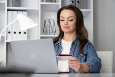 Bir kadın şık masasında oturuyor, elinde bir kredi kartı tutarken bilgisayarının ekranına odaklanıyor, online alışveriş veya modern çalışma ortamında bankacılık yapıyor.