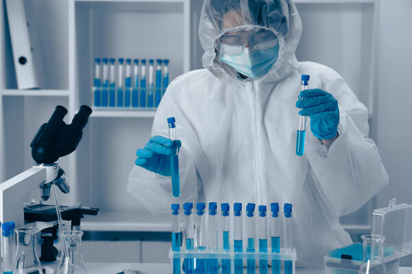 In a hightech lab, a scientist in protective gear conducts experiments, monitoring test tubes of vibrant blue liquid, with organized equipment and samples reflecting dedication to precise research