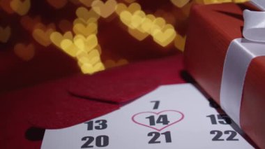14 Şubat 'ı bir kalple vurgulayan bir takvim sayfasının yakın çekimi, şenlikli bir arka plan ve güzel paketlenmiş bir hediye kutusu ile çevrili, aşkın ve kutlamanın özünü yakalıyor..