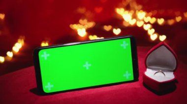 Güzel bir Sevgililer Günü sahnesinde parlak bir yüzük, kırmızı kadife bir kutunun içinde yeşil ekranlı şık bir telefonun yanında, romantik bir his için parlayan kalp ışıklarının arka planında.