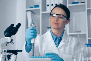 Modern bir laboratuvarda, laboratuvar önlüğü ve koruyucu gözlüklü kendini adamış bir kadın bilim adamı, gerekli laboratuvar ekipmanları ve cam parçalarıyla çevrili, hassas bir sıvıyı pipetle ustalıkla ölçer.