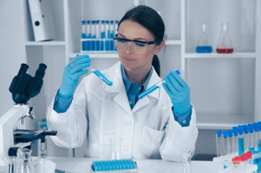 Koruyucu giysiler içinde kendini adamış bir bilimadamı, detaylı araştırma ve detaylı analizlerin derinliğini gösteren bir laboratuvar içinde çeşitli test tüplerini titizlikle inceliyor.