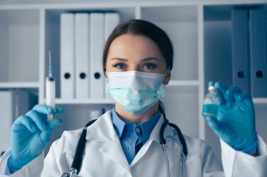 Yüz maskesi ve eldiven takmış kendini adamış bir sağlık uzmanı bir elinde şırınga, diğer elinde bir şişe, stoklanmış bir laboratuvarda duruyor ve hasta güvenliğine olan bağlılığını gösteriyor.