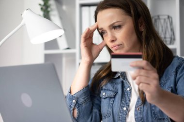 Endişeli bir kadın online alışveriş yapıyor, kredi kartını sıkıca tutuyor. Stresli görünüyor, finansal tercihlerini ve olası etkilerini düşünürken dizüstü bilgisayarına bakıyor.